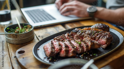 A perfectly cooked steak sits on a plate next to a laptop where a person is working, creating a delicious contrast between food and technology.