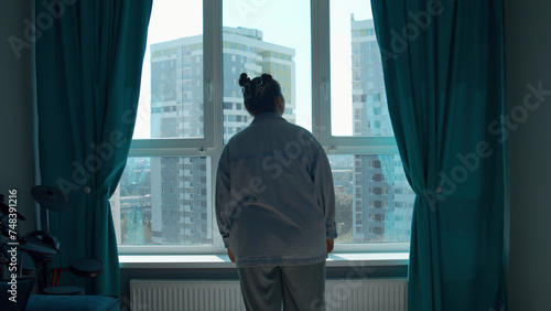 Young woman enjoying view through the panoramic window in the morning at home. Media. Girl in pajamas standing and thinking by the window in bedroom.