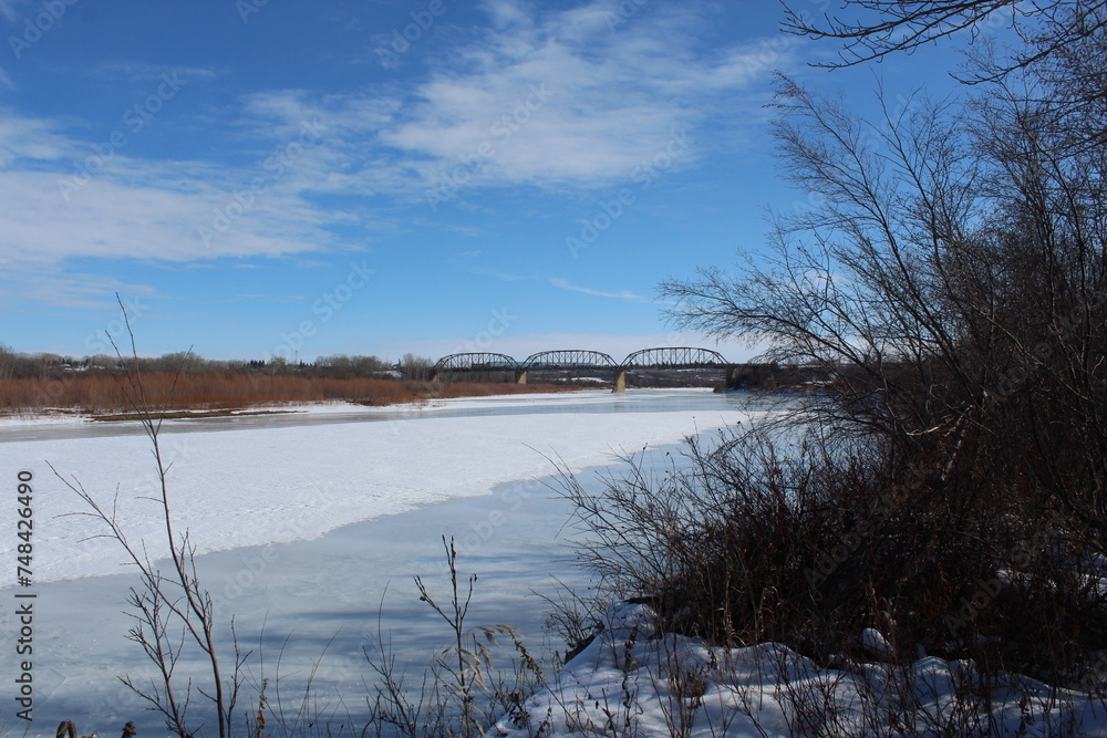 Paisaje con rio congelado y nevado con puentes al fondo y cielo azul.