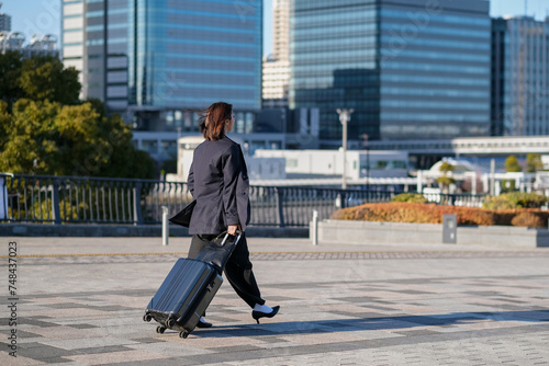 スーツケースとともに街を移動するビジネスウーマンの後ろ姿 © monzenmachi