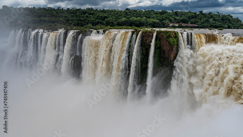 A fragment of a powerful tropical waterfall. Foaming streams of water collapse into the abyss from the ledge of the riverbed. Splashes, thick fog. green vegetation on the shore. Iguazu Falls.
