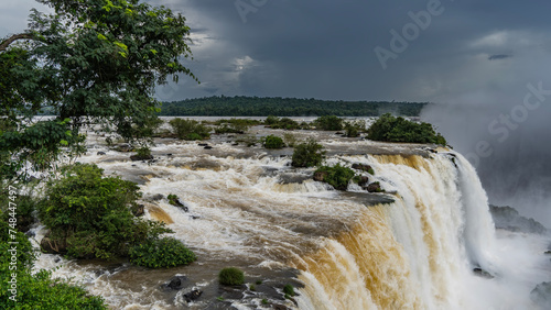 Powerful streams of white foaming water collapse from the edge of the cliff. Spray and fog rise into the sky like a cloud. Rocks with green vegetation are scattered in the stormy river. Iguazu Falls. 