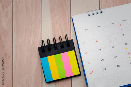 A close up of desk calendar and index sticky note on a wooden table for the concept of important dates, reminders and a well organized workspace.