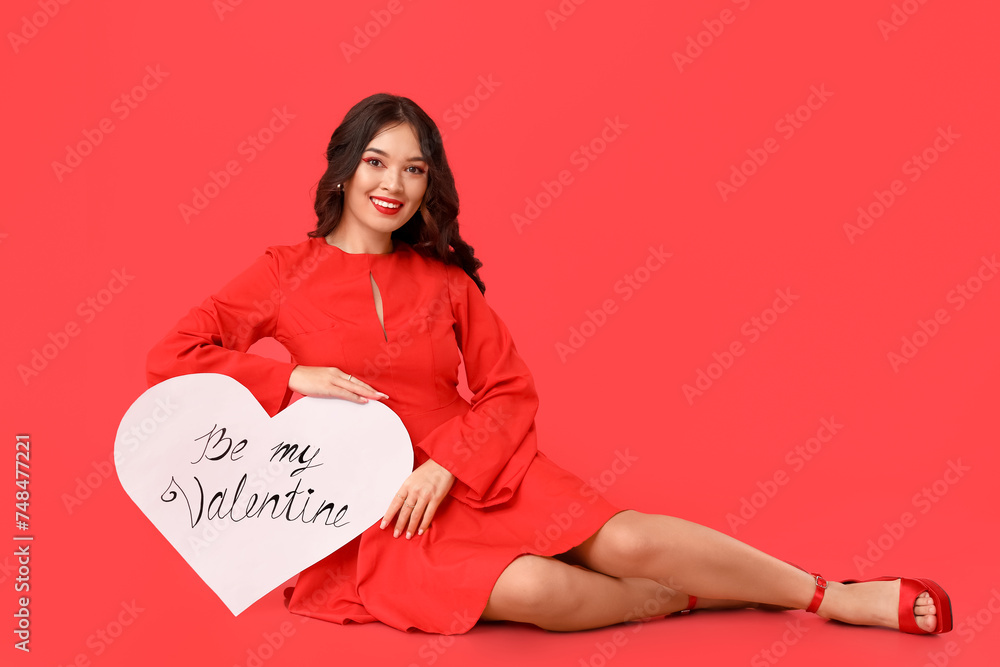 Beautiful young Asian woman with paper heart sitting on red background. Valentine's Day celebration Beautiful young Asian woman with paper heart sitting on red background. Valentine's Day celebration