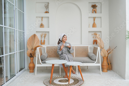 Happy attractive Asian woman in grey bathrobe posing at the terrace, smiling at camera while drinking tea. Holiday leisure concept.