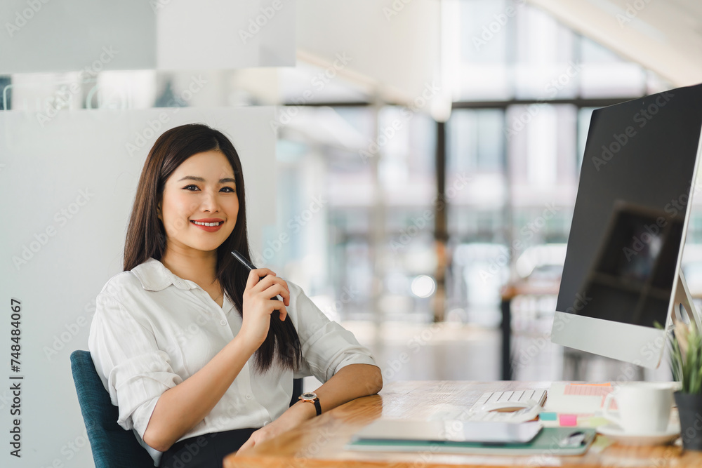 Obraz premium Smiling asian businesswoman in a casual white blouse sits confidently at her desk with a computer in a bright, modern office.