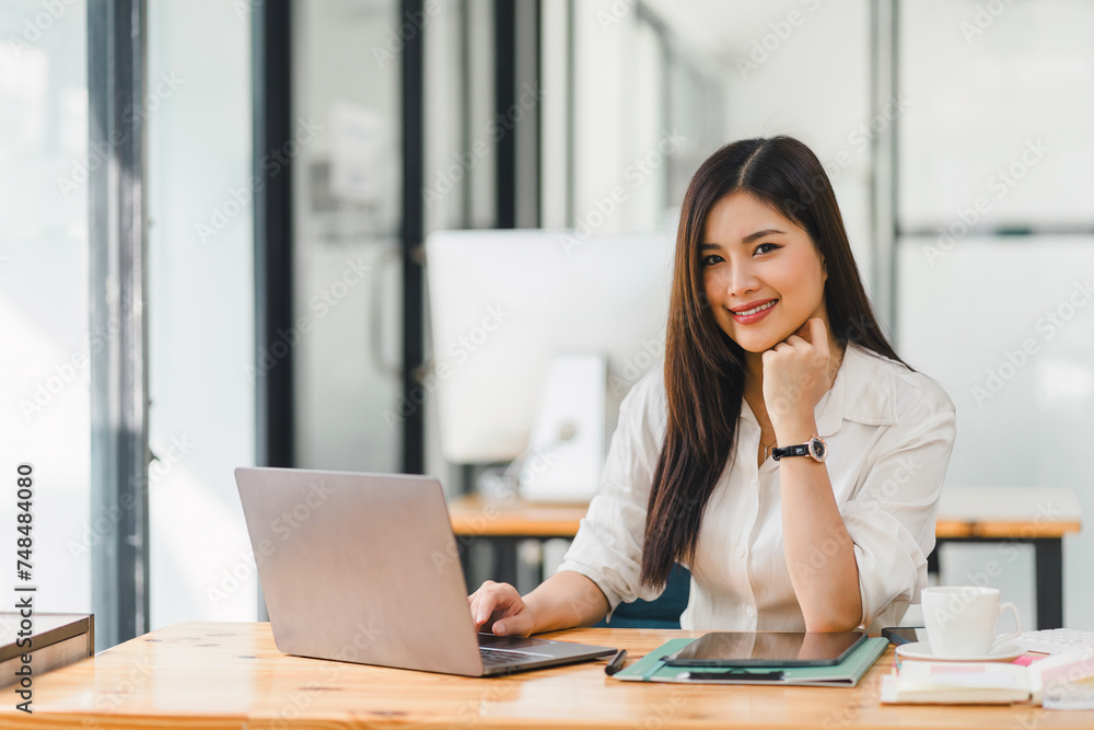 Smiling asian businesswoman in a casual white blouse sits confidently at her desk with a computer in a bright, modern office. Smiling asian businesswoman in a casual white blouse sits confidently at her desk with a computer in a bright, modern office.