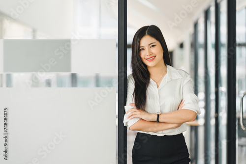 Asian professional businesswoman with crossed arms stands in a bright office corridor.