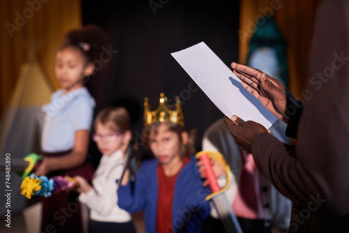 Close up of drama teacher holding play script with group of children on stage in background copy space photo