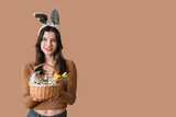 Pretty young woman in bunny ears holding wicker Easter basket with cosmetics on beige background