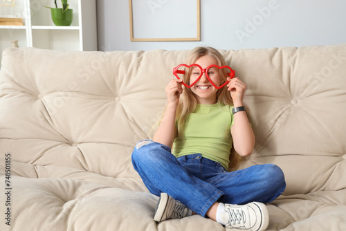 Funny little girl with big eyeglasses sitting on sofa at home. April Fools' Day celebration