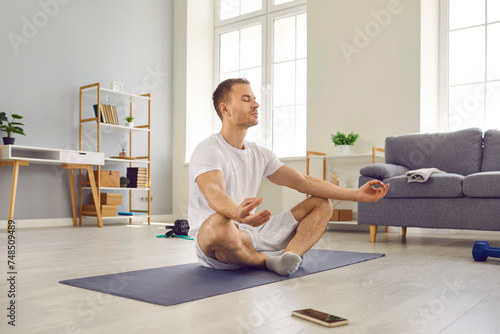Attractive man in sports outfit doing yoga and meditating on exercise mat. Sporty peaceful calm young man with closed eyes practicing yoga in lotus pose at home. Healthy lifestyle concept