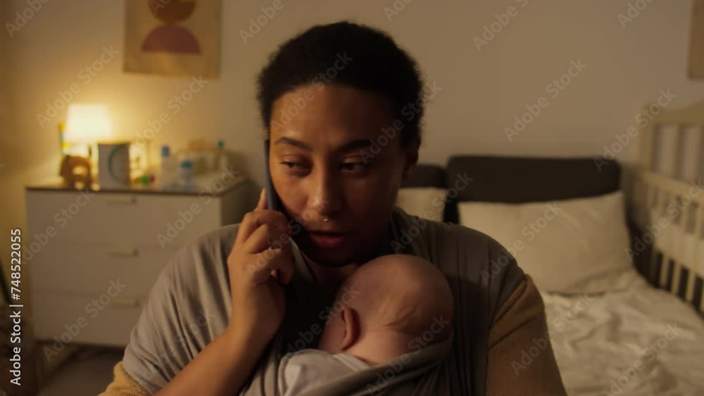 Medium close-up shot of young biracial African American woman sitting in bedroom, dandling sleeping baby son in sling wrap, and chatting on mobile phone during his daytime nap Medium close-up shot of young biracial African American woman sitting in bedroom, dandling sleeping baby son in sling wrap, and chatting on mobile phone during his daytime nap