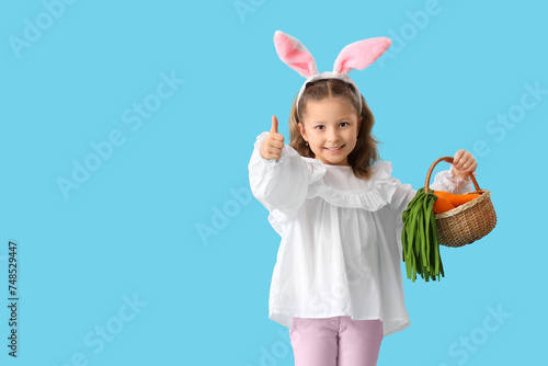 Cute little girl in bunny ears holding wicker basket with toys carrots and showing thumb-up gesture on blue background. Easter celebration