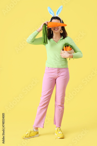 Beautiful young woman in bunny ears with toys carrots on yellow background. Easter celebration