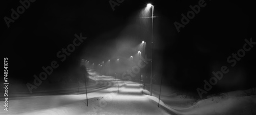 Street lights illuminate the pavement along the slippery road in a snowy winter night. 