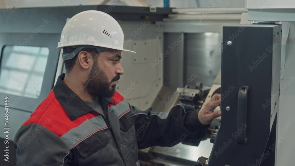 Chest up of Middle Eastern male technician wearing hard hat and workwear pressing buttons on control panel screen while setting parameters for machine equipment at manufacturing plant Chest up of Middle Eastern male technician wearing hard hat and workwear pressing buttons on control panel screen while setting parameters for machine equipment at manufacturing plant
