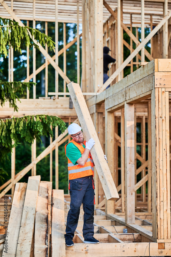 Carpenter constructs wooden-framed house near forest. Man holds large board in his hands while dressed in work clothes and helmet. Concept of modern ecological construction. Carpenter constructs wooden-framed house near forest. Man holds large board in his hands while dressed in work clothes and helmet. Concept of modern ecological construction.