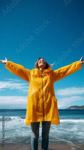 A low angle view of a happy smiling young woman wearing a yellow raincoat and standing with outstretched arms by the sea against a blue sky background. Nature  Travel  Vacations  Landscape concepts.