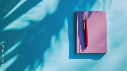 a pink notebook with a red pen on top of it next to a blue wall with a shadow of a palm tree. photo