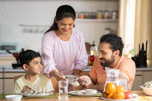 Happy Indian woman serving food to her son and husband on dining table at home - concept of family bonding, household responsibility and togetherness