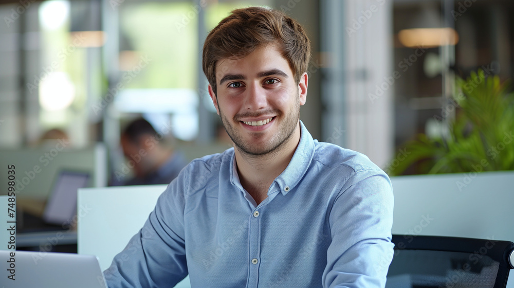 Portrait of a successful and confident young man in a blue shirt sitting in the office at a desk, working on a laptop and smiling at the camera. Portrait of a successful and confident young man in a blue shirt sitting in the office at a desk, working on a laptop and smiling at the camera.