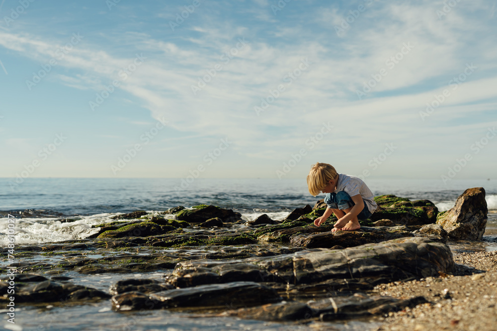 little blond boy playing on the seashore. child playing on the seashore. boy stands on stones in the water. child playing on the seashore little blond boy playing on the seashore. child playing on the seashore. boy stands on stones in the water. child playing on the seashore