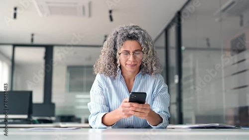 Happy middle aged business woman holding mobile cell phone using cellphone in office. Smiling mature older professional lady business owner entrepreneur using smartphone working sitting at desk. photo