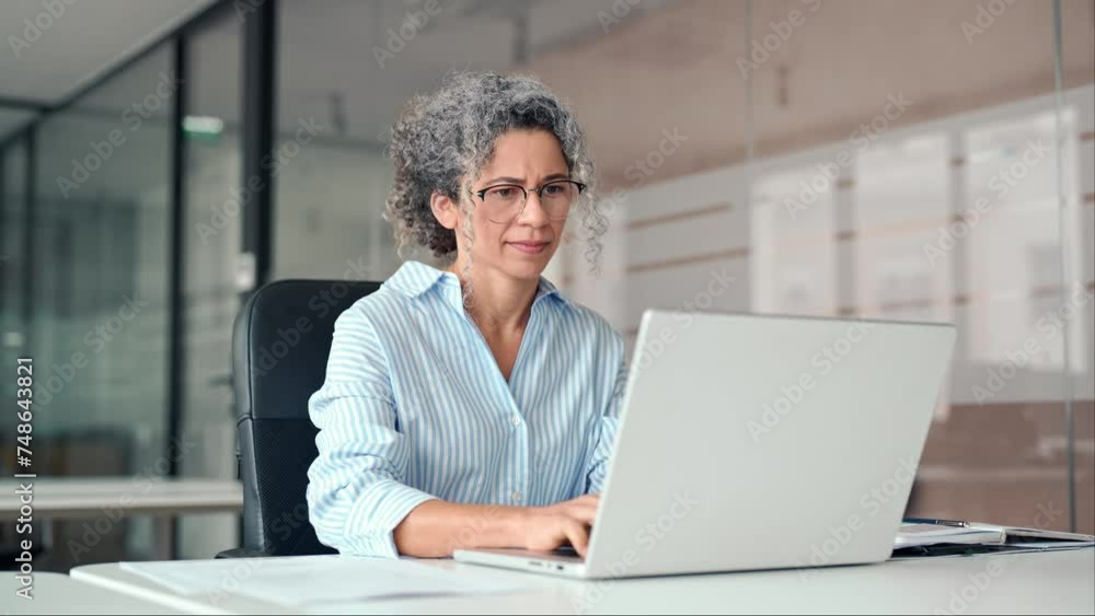 Busy older middle aged professional business woman, mature hr manager, senior lady executive leader using computer technology in modern office working looking at laptop sitting at desk.