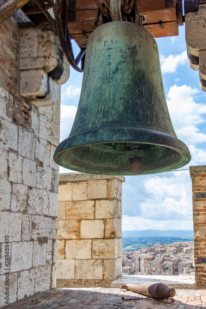 Close up view of the bell and clapper in the top of the tallest tower Torre Grossa in San Gimignano, Italy with the city buildings and Tuscan landscape seen through the crenel of wall