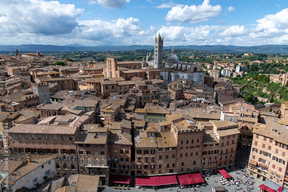 High angle view over Siena, Italy with in forefront square Piazza del Campo over the red roofs of the city toward the Siena Cathedral (Italian: Duomo di Siena) known for its facade and marble stripes