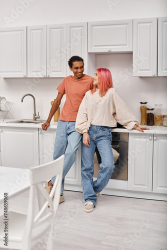 appealing multiracial joyous couple in homewear smiling happily at each other in kitchen at home