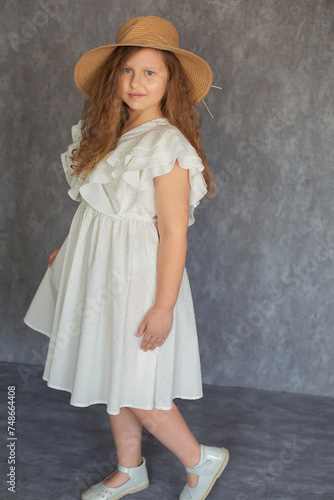 a brown-haired girl with curly hair in a white dress and a straw hat on a gray background