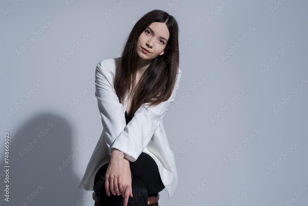 Portrait of attractive Caucasian young brunette woman in casual clothes white jacket sitting on a chair in studio. Fashion, style. Portrait of attractive Caucasian young brunette woman in casual clothes white jacket sitting on a chair in studio. Fashion, style.