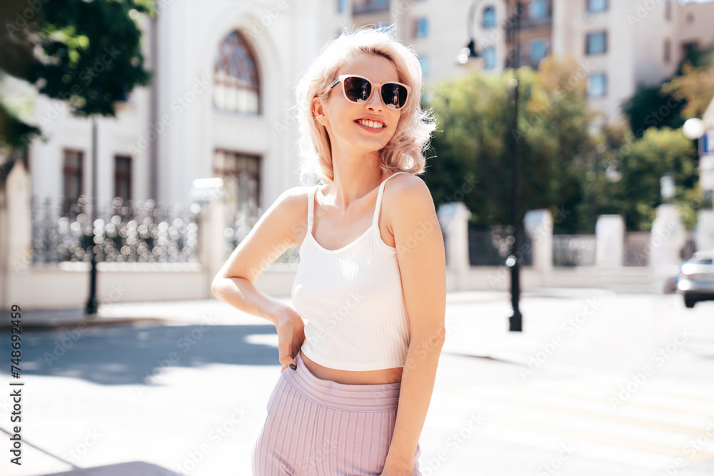 Young beautiful smiling blond hipster woman in trendy summer clothes. Carefree female posing in the street at sunny day. Positive model outdoors at sunset. Cheerful and happy sunglasses