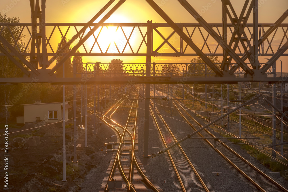 Kolorowy zachód słońca nad torami kolejowymi w Ostrowcu. Konstrukcje metalowe i tory kolejowe zanurzone w złotych promieniach zachodzącego słońca w majowy wieczór. - obrazy, fototapety, plakaty 