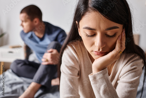 young and offended asian woman with closed eyes near husband sitting on blurred background on bed photo