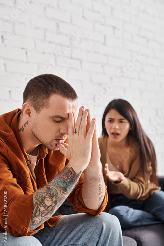 young tattooed man sitting with closed eyes near angry asian man quarrelling at home, divorce photo