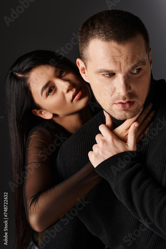 seductive asian woman embracing young man in black outfit and looking at camera on grey backdrop