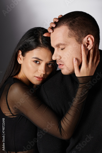 brunette passionate asian woman in black casual attire embracing head of stylish man on grey