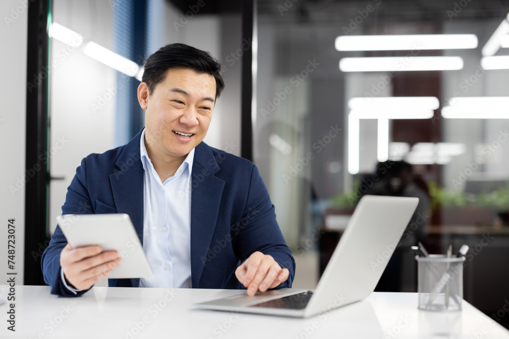 Modern businessman in formal clothes using digital tablet while working by pc on white table. Successful entrepreneur using two wireless devices for comparing articles from different web sources. Modern businessman in formal clothes using digital tablet while working by pc on white table. Successful entrepreneur using two wireless devices for comparing articles from different web sources.