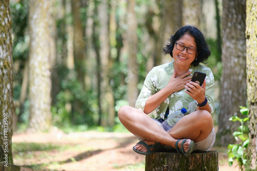 A woman sitting on a wood beam is holding a smartphone; smiling, happy face photo