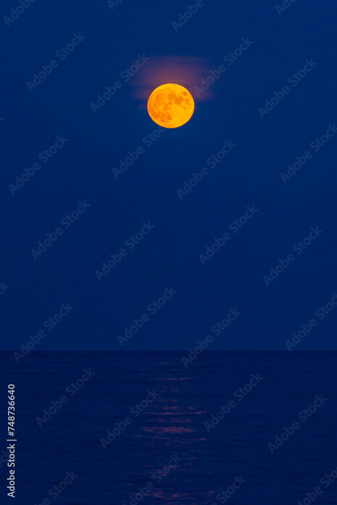 The yellow moon rises above the blue water and sky. High quality photo