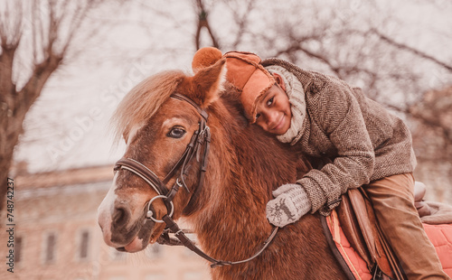 a boy sitting in a saddle lies on the neck of a pony and smiles looking to the side © Anna