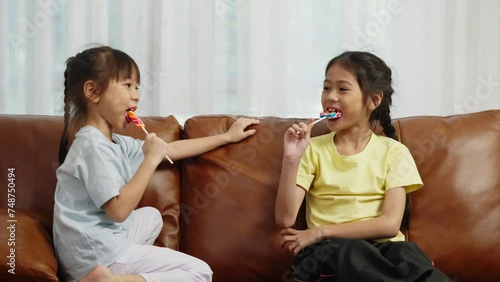 Footage handheld medium shot, Two cute girls sitting on sofa and enjoy to play and eating big lollipop together, Asian sibling girls talking with fun in living room at home