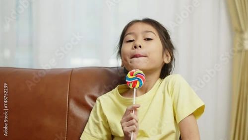 Footage handheld wide shot, Two cute girls sitting on sofa and enjoy to play and eating big lollipop together, Asian sibling girls talking with fun in living room at home