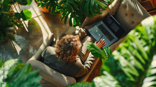 Busy elegant mature middle aged business woman professional leader wearing suit holding laptop using computer working sitting in comfortable chair in sunny office with green plants. Overhead top view.
