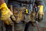 Close-up of a worker's tool belt with various tools and yellow gloves.