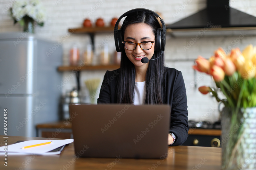 An Asian woman, smiling, communicates with a headset on, providing professional online support from her home office. An Asian woman, smiling, communicates with a headset on, providing professional online support from her home office.