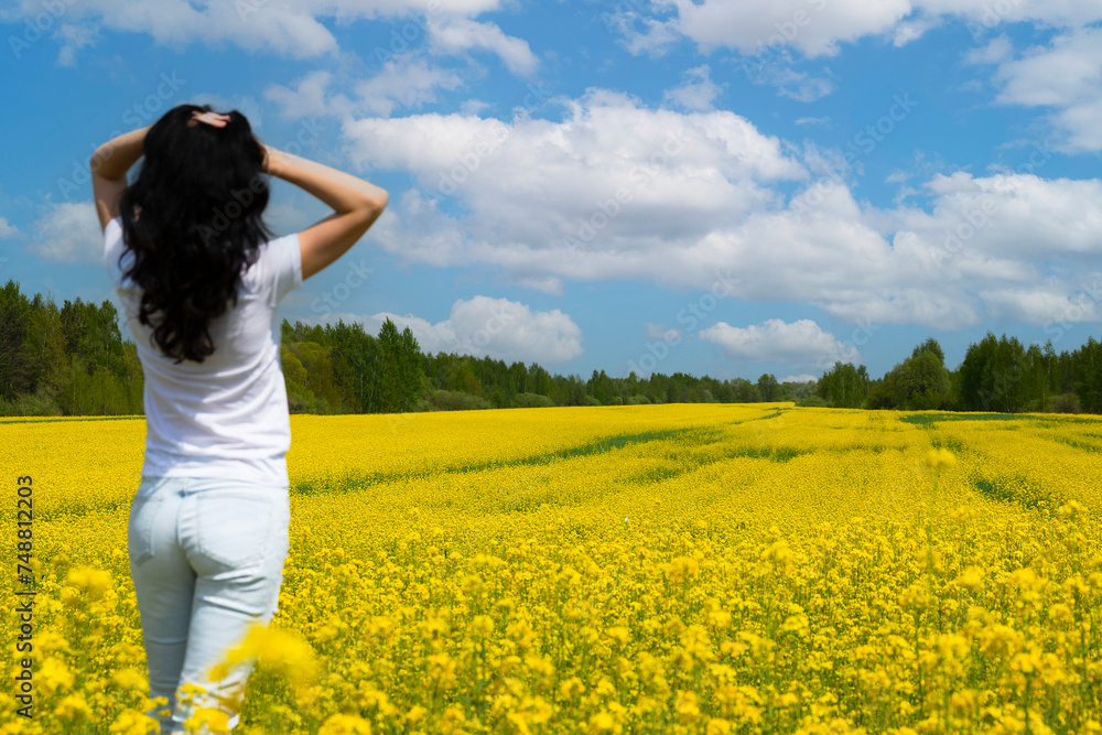 Rapeseed field with yellow flowers, blue sky with clouds and dark-haired beautiful brunette girl, view from the back. Copy space. Rapeseed field with yellow flowers, blue sky with clouds and dark-haired beautiful brunette girl, view from the back. Copy space.